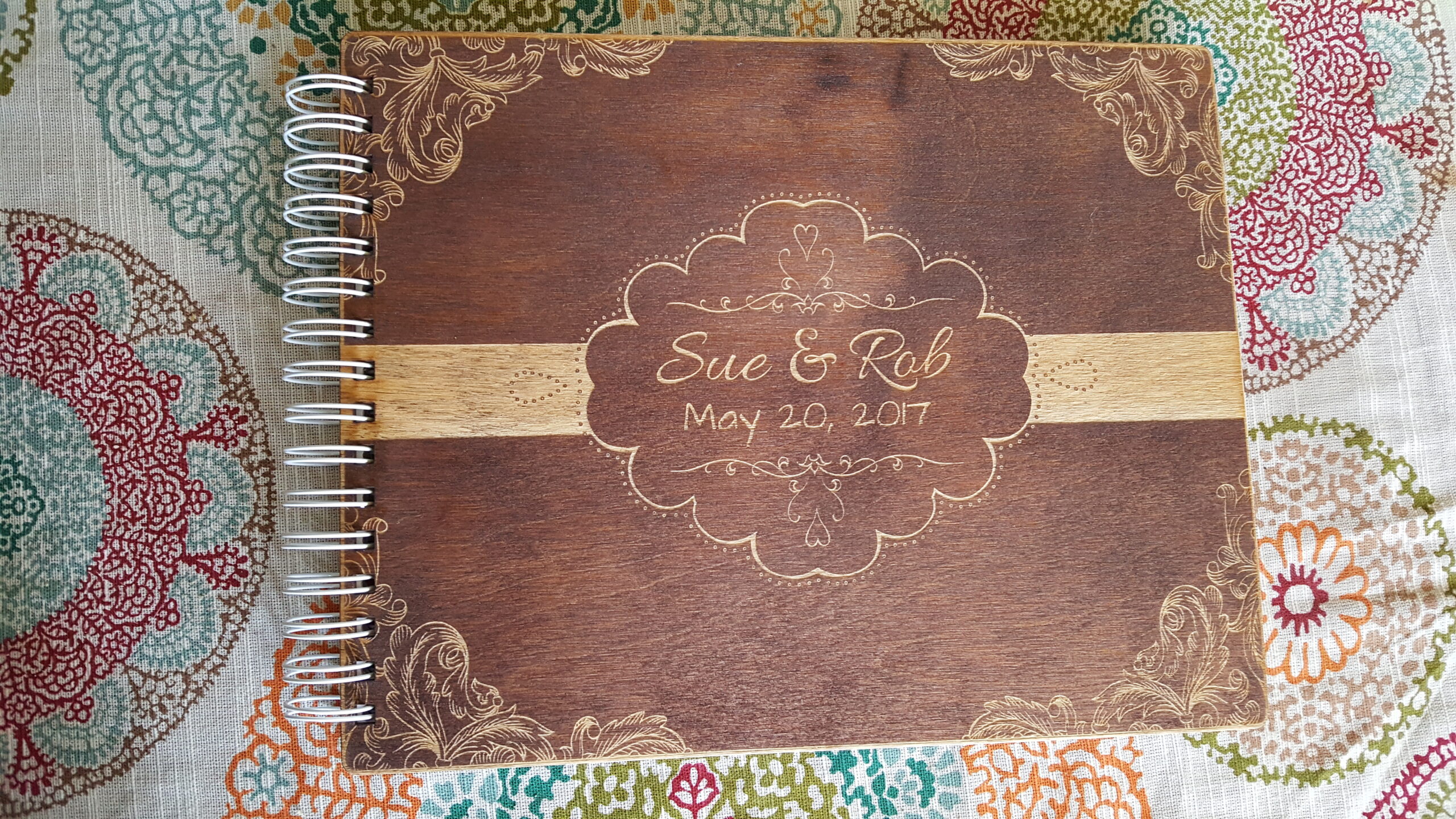 Wooden spiral-bound notebook with engraved names and date on a patterned fabric.